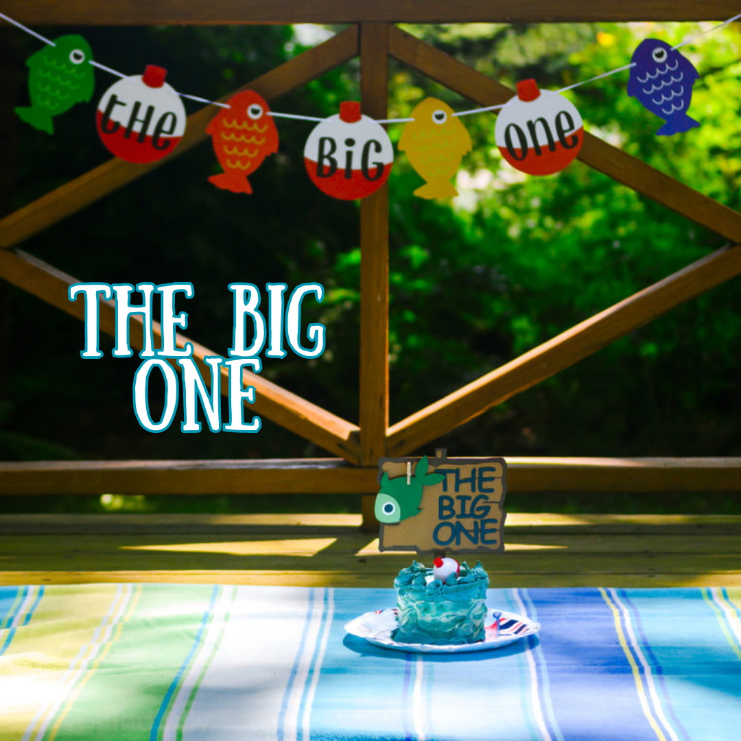 A blue, water and fish themed smash cake sits against brown wood and green trees in the background. Text Reads 